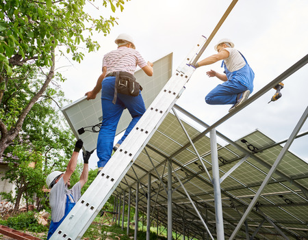 Installing of stand-alone solar photo voltaic panel system. Team of three technicians mounting big shiny solar module on high metal platform. Alternative energy and professional construction concept.の写真素材
