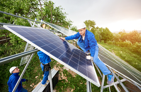 Installing of stand-alone solar photo voltaic panel system. Two technicians in hard-hats mounting big shiny solar module on platform on green summer view background. Alternative energy concept.の写真素材