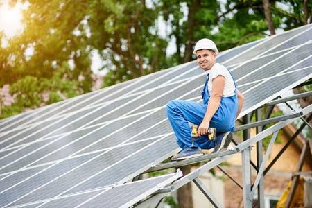Young smiling electrician worker sitting on almost finished stand-alone solar photo voltaic panel system on bright sunny green tree background. Alternative energy concept.の写真素材