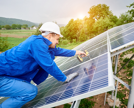 Construction worker in hard hat and blue overall connects photo voltaic panel to solar system using screwdriver. Alternative cheap sun energy production and profitable financial investment concept.の写真素材