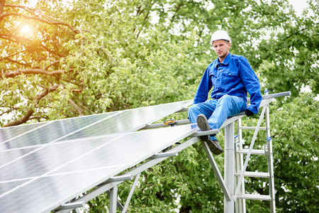 Young smiling electrician points screwdriver at camera sitting on almost finished stand-alone solar photo voltaic panel system on bright sunny green tree background. Alternative energy concept.の写真素材