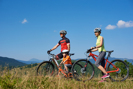 Young family tourist bikers, mom, dad and child resting with bicycles, looking into distance, standing on grassy hill on summer sunny day. Active lifestyle, traveling and happy relations concept.の写真素材