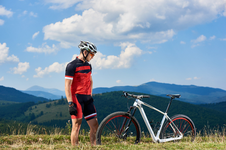 Young athletic sportsman biker in professional sportswear looking at mtb on mountain grassy hill on bright blue summer sky background. Tourism, active lifestyle and extreme sport concept.の写真素材