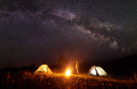 Camping in mountains. Young slim tourist girl standing between two tents watching brightly burning bonfire on deep dark sky with lot of bright sparkling stars background. Tourism and travel concept.の写真素材