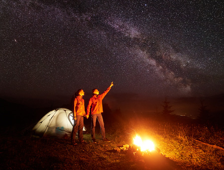 Night camping in mountains. Couple standing in front of illuminated tent, holding hands lit by burning campfire, man pointing at starry sky and Milky way to woman. Tourism and beauty of nature conceptの写真素材