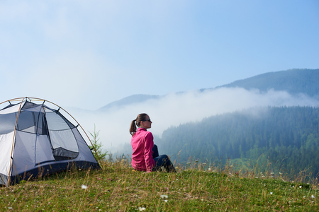 Back view of young long-haired tourist girl in sunglasses sitting on green grass of blooming valley at tourist tent under beautiful clear blue sky enjoying mountains view on bright summer morning.の写真素材