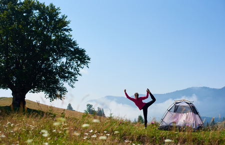 Back view of young slim girl standing on one leg in yoga pose on green grassy hill at tourist tent and big tree under beautiful blue sky on foggy mountains background. Tourism and camping concept.の写真素材