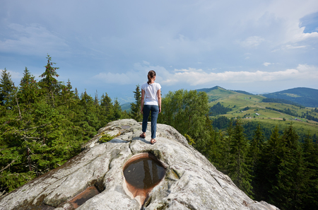 Back view of young long-haired tourist girl standing on huge rock with big puddle in the middle under beautiful blue sky enjoying fantastic view of mountains covered with ever-green pine forest.の写真素材