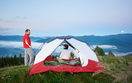 Man standing near the tent in which sits a woman against the beautiful scenery of the mighty mountains in the fog under the morning sky on which the moon is visible in the distance.の写真素材