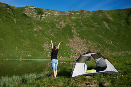 Back view of young woman tourist, standing with lifting hands up in the air near tent, enjoying sunny day near the lake in the mountains. Lifestyle concept adventure summer vacations outdoorの写真素材