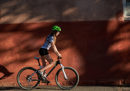 Young sporty woman in cycling clothes and helmet riding bike in front of red wall. Concept of healthy lifestyle and everyday activities. Female exercising, overcoming challanges, morning routine.の写真素材