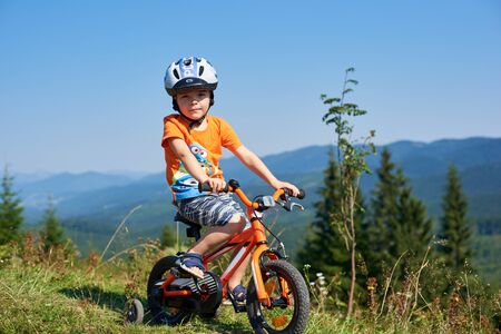 Verkhovyna, Ukraine - August 19, 2017: Photo of small cute preschool boy in helmet, shorts and T-shirt looking in camera cycling three-wheeled child bike on grassy hill on beautiful warm sunny day.のeditorial素材