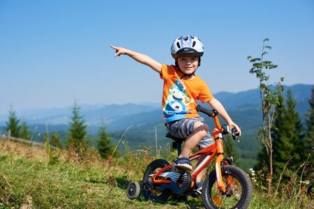 Verkhovyna, Ukraine - August 19, 2017: Small preschool boy in helmet and summer clothes points at something in distance riding child bike down steep hill smiling in camera on mountains background.のeditorial素材