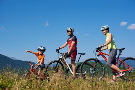 Verkhovyna, Ukraine - August 19, 2017: Small boy in helmet points his mom and dad at something in distance. Young tourist family riding bikes in high grass on mountain background on warm summer day.のeditorial素材