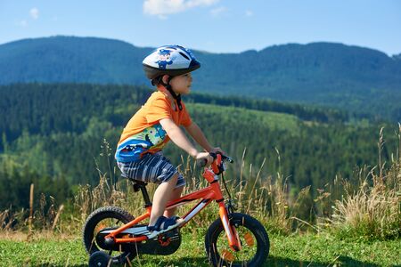 Verkhovyna, Ukraine - August 19, 2017: Small cute preschool boy in helmet, shorts and T-shirt cycles three-wheeled child bike on grassy valley on beautiful green mountains and blue sky background.のeditorial素材