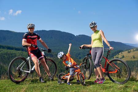 Verkhovyna, Ukraine - August 19, 2017: Young tourist family bikers, mother, father and small funny boy leaning on bicycles enjoying rest on grassy valley under blue sky on distant mountains backgroundのeditorial素材