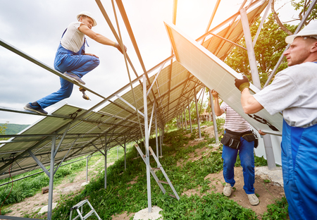 Installing of solar photo voltaic panel system. Three technicians lifting heavy solar module on high platform. Investment in alternative energy, money saving and professional construction concept.の写真素材