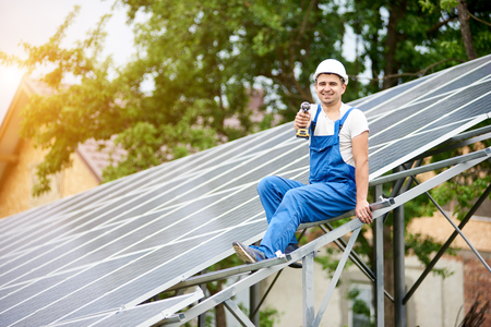Young smiling electrician points screwdriver at camera sitting on almost finished stand-alone solar photo voltaic panel system on bright sunny green tree background. Alternative energy concept.の写真素材
