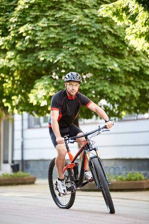 Young male bicyclist in cycling sportswear and protective helmet, looking at camera, riding bike down empty city center alley surrounded by green trees. Sportsman training outdoors improving skills.の写真素材