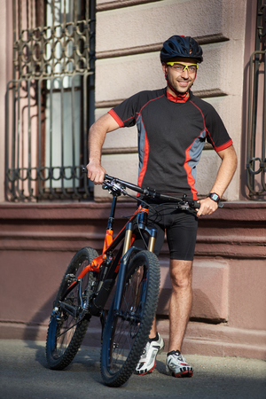 Young male biker in cycling clothes and protective helmet standing near old historical building next to bicycle looking far and smiling. Sportsman taking break after bicycle training in city center.の写真素材
