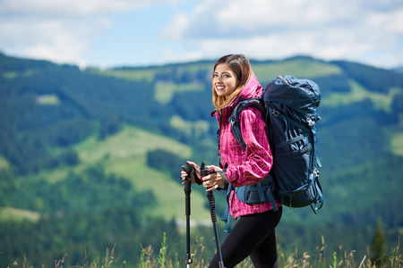 Close-up portrait of smiling active female backpacker with backpack and trekking poles, wearing sports wear, travelling, looking away, enjoying sunny day in the mountains. Copy spaceの写真素材