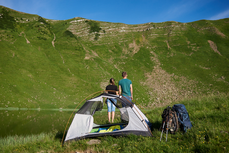 Rear view of a guy and a girl stand near camping with a tent, backpacks and trekking sticks, looking at a mighty green mountain at the foot of which is a lake under a blue skyの写真素材