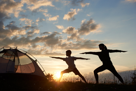 Silhouette view of Yoga Warrior pose exercising by family couple in grass near tent at the daybreak on the background of morning sky with sparse clouds and bright sun. Mom grafts a sons love of sportの写真素材