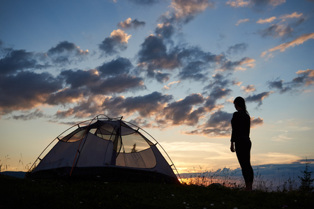 Back view of silhouette of girl backpacker standing on top of mountain near camping on a grass with wildflowers at daybreak enjoying beautiful scenery of sunset in the evening sky in the rare clouds.の写真素材