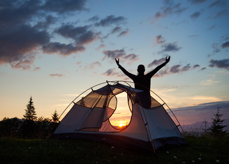 Silhouette of a tourist female standing with her hands raised to the sky on top of a mountain near campsite at sunset. Through tent visible bright sun over the mountains. Rear viewの写真素材