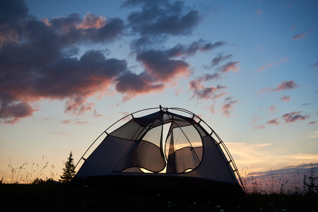 Camping at sunrise in the mountains. The tent stands on the grass with wildflowers under the morning sky with clouds on top of the mountain. The concept of an active lifestyleの写真素材
