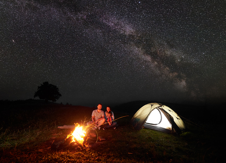 Modern tourist family camping in mountains by night. Husband and wife with small daughter sitting on a log in front of tent at burning campfire and look in camera. Tourism and traveling concept.の写真素材