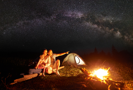 Camping in mountains at night. Tourist family resting in front of illuminated tent and burning bonfire. Woman holding in arms small girl, man pointing at bright stars in dark sky and Milky wayの写真素材