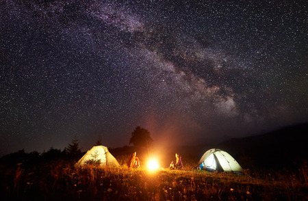 Night camping in mountains. Bright campfire burning between two backpackers, man and woman sitting opposite each other in front of illuminated tents under amazing dark blue starry sky and Milky wayの写真素材