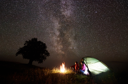 Romantic couple man and woman resting near glowing tent, bonfire and silhouette of big tree under starry sky and Milky way on background, enjoying night camping. Tourism, outdoor activity conceptの写真素材