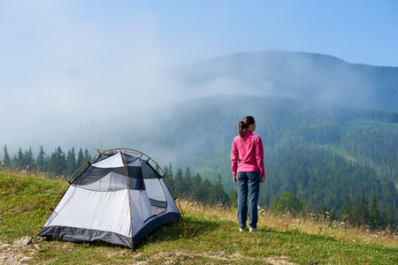 Back view of young tourist woman standing on blooming valley at tourist tent under beautiful clear blue sky, enjoying foggy mountains view on summer morning. Tourism and traveling concept.の写真素材