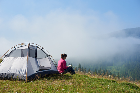 Back view of young female backpacker sitting on green grass of blooming valley at tourist tent under beautiful blue sky reading a book on bright summer morning on foggy mountains background.の写真素材