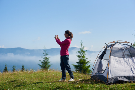 Happy tourist girl standing on blooming grassy valley in front of small tourist tent and taking picture of beautiful mountains covered with white clouds under clear blue sky on bright summer morning.の写真素材