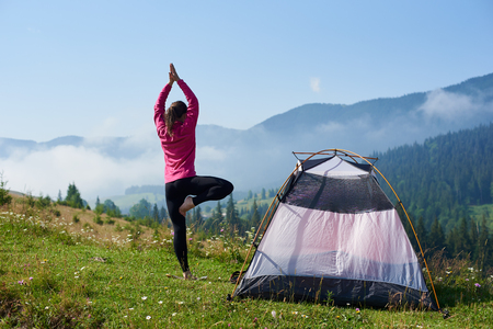 Back view of young long-haired barefooted tourist girl standing on one leg in yoga pose with raised arms on green grassy valley at tourist tent under beautiful blue sky on foggy mountains background.の写真素材