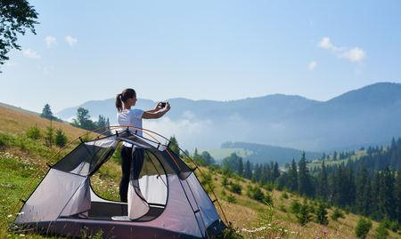 Young pretty slim girl standing on steep grassy hill at small tourist tent and taking picture of beautiful foggy mountains covered with forest under clear blue sky on bright summer morning.の写真素材