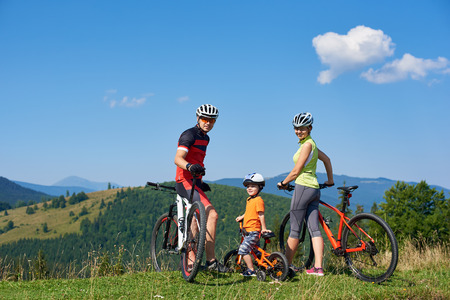 Young happy family tourists bikers, mom, dad and child resting with bicycles on grassy hill, looking in camera. Mountains view and blue sky on background. Healthy lifestyle and relations concept.