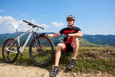 Professional sportsman cyclist in sportswear, helmet sitting near his bicycle on grassy roadside, resting after cycling bike in mountains on sunny summer day. Active lifestyle, outdoor sport concept.の写真素材