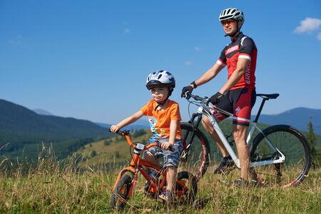 Verkhovyna, Ukraine - August 19, 2017: Tourist bikers, dad and small boy in helmets and bright clothes on mountain bicycles in high grass on beautiful sky background. Tourism and traveling concept.のeditorial素材