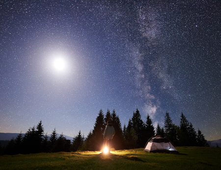 Camping site in mountain valley at night. Male hiker standing near tourist tent at campfire under night blue starry sky with Milky way and bright full moon. Outdoor activity and traveling conceptの写真素材