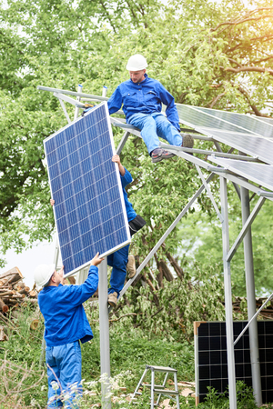 Installing of stand-alone solar photo voltaic panel system. Workers in hard-hats and blue overall lifting the solar module on metal platform. Alternative energy and professional construction concept.の写真素材