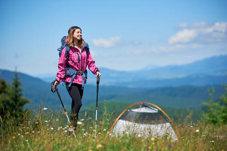 Sporty smiling female traveller with backpack and trekking sticks near tent, wearing sports wear, standing on the top of a hill against blue sky and clouds, enjoying sunny day in the mountainsの写真素材
