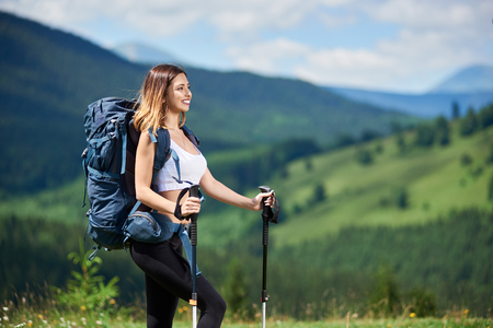 Close-up portrait of beautiful sporty woman traveller with blue backpack and trekking sticks, hiking on the top of a hill, looking away, enjoying sunny day in the mountains. Copy spaceの写真素材