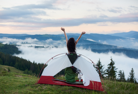 Back view of woman camper standing in the tent entrance in sleeping bag, lifting hands up in the air, enjoying beautiful view of foggy valley, mountains in the morning. Camping lifestyle conceptの写真素材