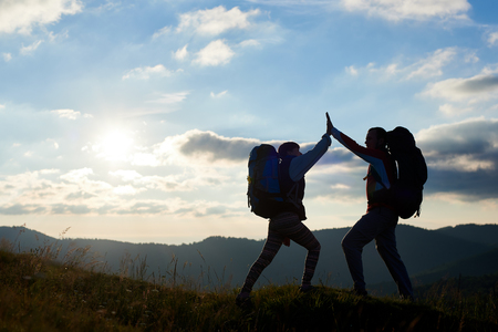 Silhouette of happy couple with backpacks on top of the mountain give each other a high five against the background of the mountains and the cloudy sky with a bright sun at sunset. Bottom viewの写真素材