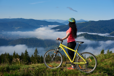 Back view of sexy woman cyclist with yellow bicycle on a rural trail in the mountains, wearing helmet and red red t-shirt, enjoying morning haze in valley, forests on the blurred backgroundの写真素材