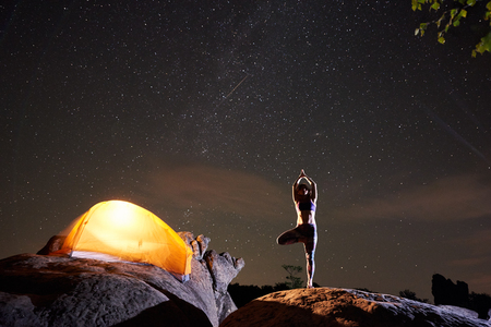 Brightly lit tourist tent and young slim woman standing on one leg with raised arms doing yoga at night on huge boulder under dark starry sky. Tourism, nature and active lifestyle concept. Vrikshasanaの写真素材
